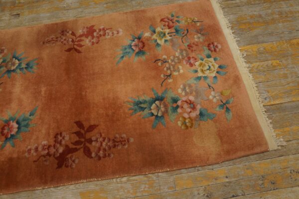 Pink-orange rug featuring colorful floral motifs rests on worn, yellow-toned wooden floors.