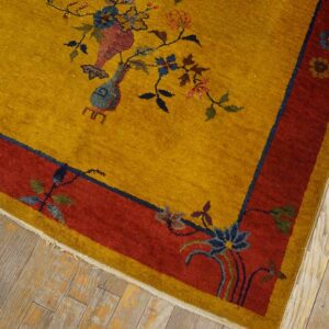 Gold high-pile rug with red borders and colorful floral vases rests on light, distressed wood plank flooring.