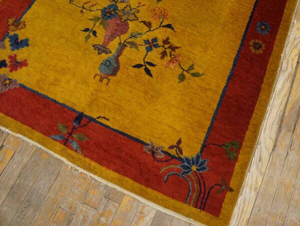 Gold high-pile rug with red borders and colorful floral vases rests on light, distressed wood plank flooring.