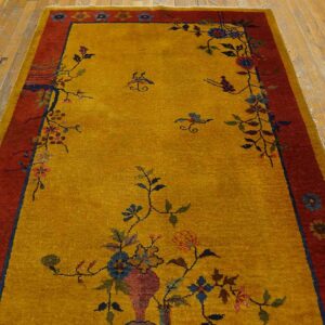Gold field rug with multicolored floral designs and a wide red border on light wood planks.