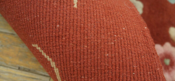 Detail view of a dense, rust-colored rug surface with subtle cream geometric and blurred pink patterns.