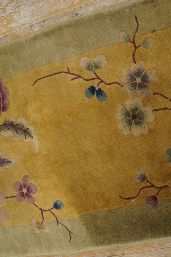 Gold rug with a sage green border and stylized floral pattern rests on distressed pale wood planks.