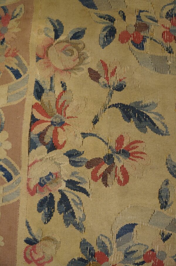 Worn flat-weave rug featuring faded blue and red floral patterns on a tan ground with a rose border.