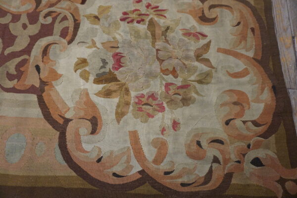 Ornate flatweave rug displaying a faded floral medallion and curving border motifs in beige, rose, and rust.