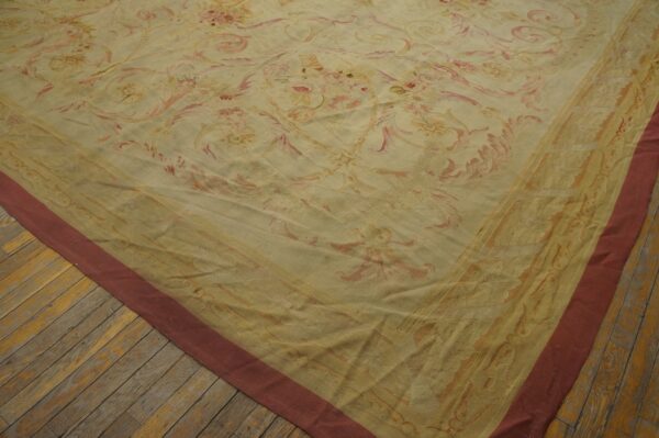 Corner of a yellow and pink ornate floral rug with a reddish border rests on distressed wood planks.