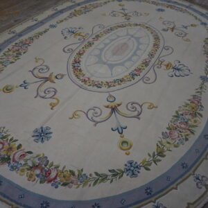 Oval cream rug with a pastel floral medallion and blue border, lying on distressed wood planks.