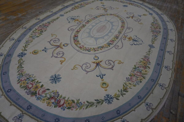 Oval cream rug with a pastel floral medallion and blue border, lying on distressed wood planks.