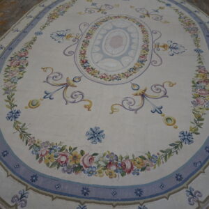 Oval cream rug featuring a central blue medallion and borders filled with colorful floral garlands on wood planks.