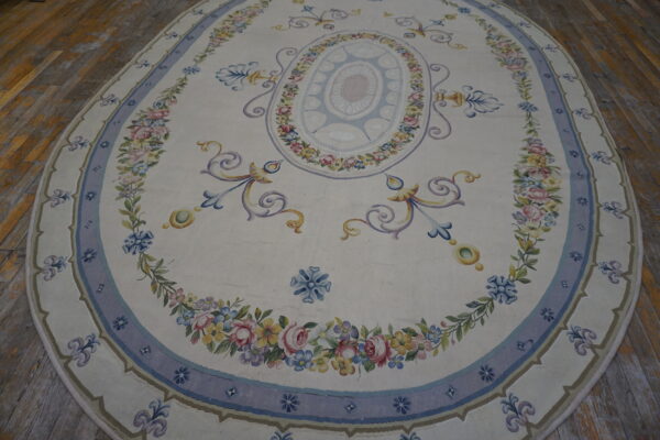 Oval cream rug featuring a central blue medallion and borders filled with colorful floral garlands on wood planks.