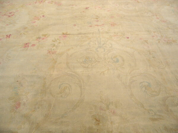 Muted beige rug surface with faint pink floral designs and low-contrast blue classical scrolling patterns.