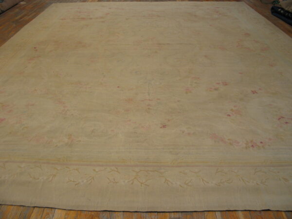 Tan low-pile rug with a low-contrast floral pattern and border, displayed on hardwood flooring.