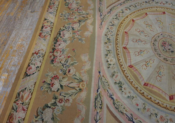 Close-up of a pastel rug featuring floral garlands, pink stripes, and a round medallion on distressed plank flooring.