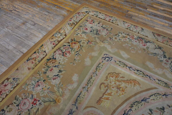 A low-pile tan rug with elaborate pink and cream floral borders lies on distressed grey wooden floors.