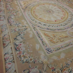 Neoclassical rug in tan, pale pink, and cream with a detailed floral border rests on distressed plank flooring.