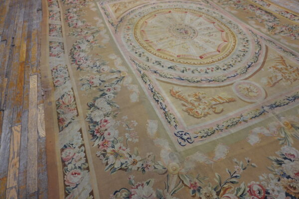 Neoclassical rug in tan, pale pink, and cream with a detailed floral border rests on distressed plank flooring.