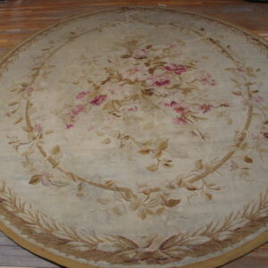 Muted beige round rug with a central pink floral pattern and tan scrolling vines on wood planks.