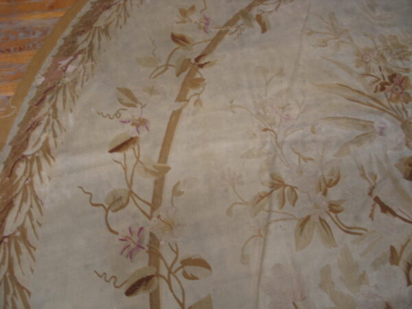 Muted tan rug with a floral vine pattern, showing a decorative leaf border over wood flooring.