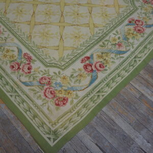 Cream rug with a pink rose and blue ribbon floral border rests on distressed gray wooden flooring.