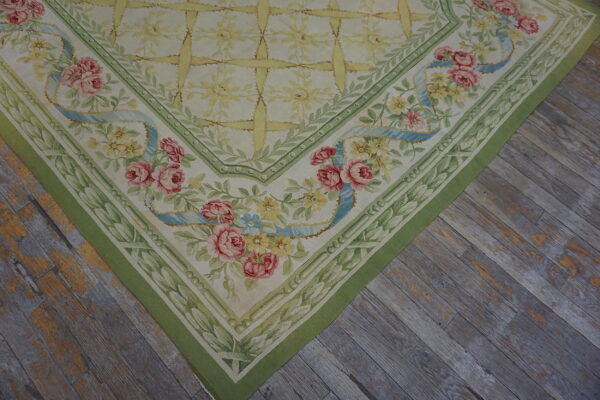 Cream rug with a pink rose and blue ribbon floral border rests on distressed gray wooden flooring.