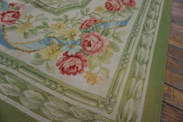 Flatweave rug corner with pink roses, yellow florals, and sage green borders on rustic wood planks.