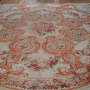 Large ornate rug featuring faded coral scrollwork and pink florals on a pale background over wood flooring.
