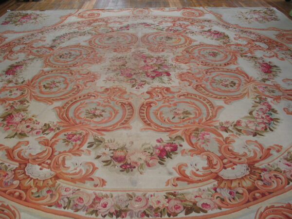 Large ornate rug featuring faded coral scrollwork and pink florals on a pale background over wood flooring.