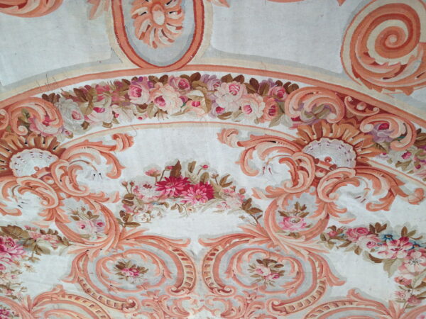 Ornate low-pile rug with salmon neoclassical scrolls and multi-colored pink floral bouquets on a pale background.