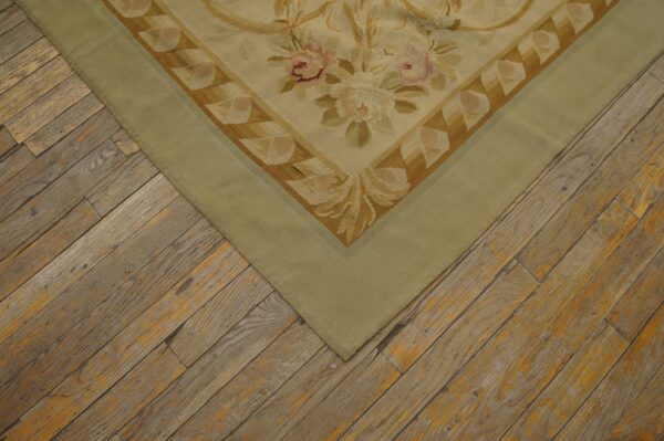 Flatweave rug corner featuring beige, pink, and olive floral motifs with a wide sage green border on weathered wood.