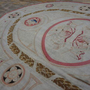 Round low-pile rug with a classical pattern in pale pink, brown, and white over worn hardwood.