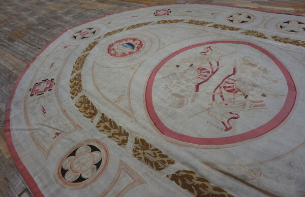 Round low-pile rug with a classical pattern in pale pink, brown, and white over worn hardwood.