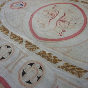A close-up view of a faded, flat-woven rug featuring classical medallions, vine borders, and rose and brown accents on a pale ground.