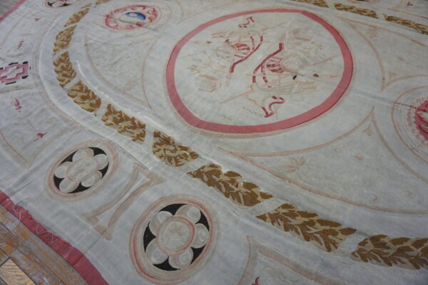 A close-up view of a faded, flat-woven rug featuring classical medallions, vine borders, and rose and brown accents on a pale ground.