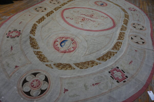 Large oval white rug with pink, tan, and blue ornate motifs and laurel wreaths on a distressed wood floor.