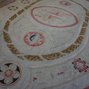Pale oval rug with a central pink medallion, brown leaf wreaths, and geometric motifs on a wood floor.