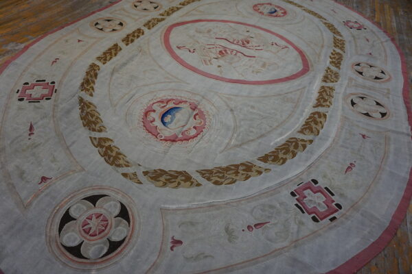 Pale oval rug with a central pink medallion, brown leaf wreaths, and geometric motifs on a wood floor.