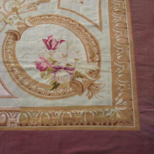 Classical style rug with pink florals, tan scrollwork, and a maroon border visible over wood flooring.