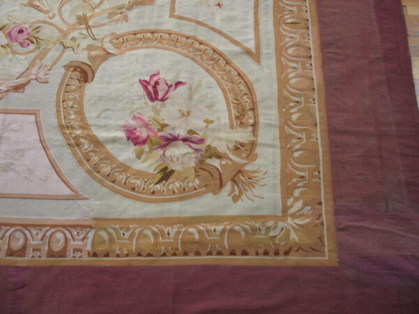 Classical style rug with pink florals, tan scrollwork, and a maroon border visible over wood flooring.