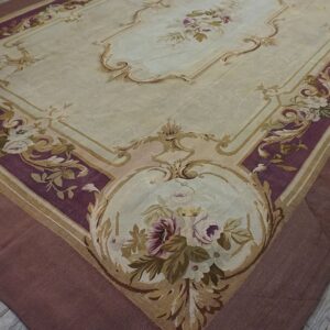 Low pile rug with light beige field and ornate plum and gold scrolling floral borders on wood flooring.