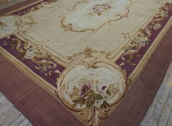 Low pile rug with light beige field and ornate plum and gold scrolling floral borders on wood flooring.