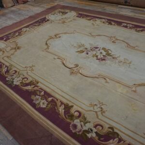Ornate light beige rug with deep purple borders and scrolling floral designs lies on distressed wood plank flooring.