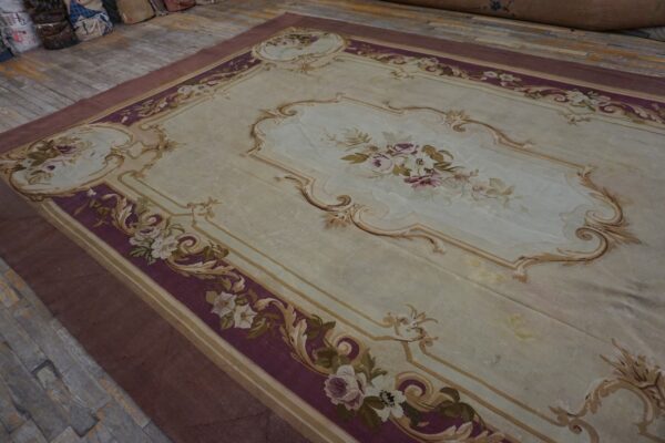 Ornate light beige rug with deep purple borders and scrolling floral designs lies on distressed wood plank flooring.