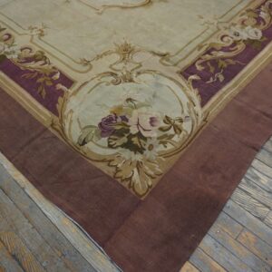 Ornate low-pile rug with cream, purple, and gold floral patterns bordered in reddish-brown, on distressed wood floorboards.