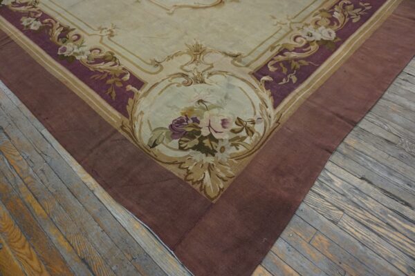 Ornate low-pile rug with cream, purple, and gold floral patterns bordered in reddish-brown, on distressed wood floorboards.