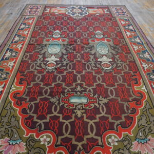 Ornate low-pile rug with a maroon and red geometric field, colorful borders, placed on distressed wood planking.