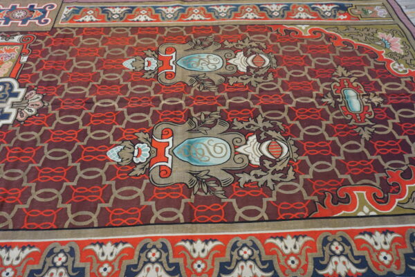 Ornate low-pile rug with maroon, red, gold, and blue motifs over a geometric chain pattern.