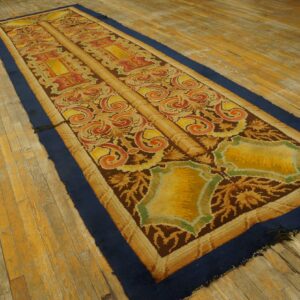 Low-pile runner rug with scrolling brown, yellow, and orange motifs, bordered in deep blue, on wood planks.