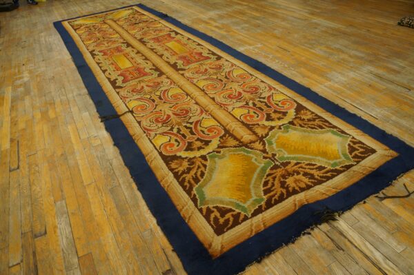 Low-pile runner rug with scrolling brown, yellow, and orange motifs, bordered in deep blue, on wood planks.