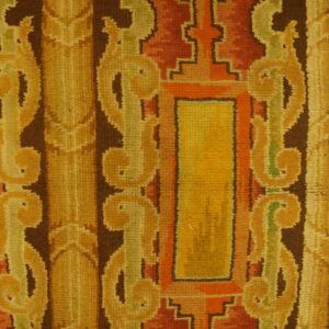 Medium-pile rug detail showing gold and rust geometric patterns, dark brown accents, a navy border, and wood flooring.