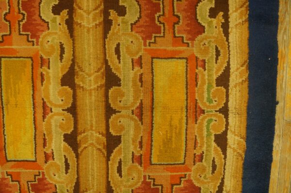 Medium-pile rug detail showing gold and rust geometric patterns, dark brown accents, a navy border, and wood flooring.