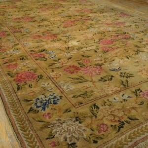 Large gold floral rug with pink and blue motifs features a fringed edge on light wood planks.
